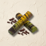 A 300x300 square professional food photograph featuring two cylindrical gourmet Rum Runner truffle bars. The bars are arranged in an "X" shape and feature a vibrant, hand-painted finish with a marbled blend of bright yellow and mossy green, decorated with bold, horizontal dark chocolate stripes. The bars rest on a heavily textured, light cream-colored plaster surface. Scattered around the base are delicate dark chocolate curls, enhancing the artisanal presentation. The lighting is bright and clean, emphasizing the unique colors and textures of the truffles.