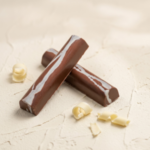 A 300x300 square professional food photograph of two cylindrical gourmet Sea Salt Caramel truffle bars. The bars are made of smooth milk chocolate and feature thick, artistic white chocolate swirls running lengthwise. They are arranged in an "X" shape on a textured, light cream-colored plaster surface. Several delicate white chocolate curls are artfully scattered around the base of the truffles. The lighting is bright and clean, emphasizing the glossy finish of the chocolate and the sophisticated, artisanal aesthetic.