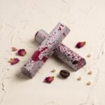 A 300x300 square professional food photograph featuring two cylindrical gourmet Sparkling Shiraz truffle bars. The bars are arranged in an "X" shape and feature a unique artisanal finish: a matte white base heavily speckled with fine dark purple dots and bold, irregular splatters of deep burgundy wine-colored glaze. The text "SPARKLING SHIRAZ" is centered at the bottom in a sophisticated, muted rose-toned serif font. The bars rest on a heavily textured, light cream plaster surface. Elegant decorative accents include scattered dried pink rose petals, a single roasted coffee bean, and delicate white decorative pearls. The lighting is soft and bright, creating a premium, romantic, and curated aesthetic.