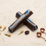A 300x300 square professional food photograph featuring two cylindrical gourmet "Spiked Joe" truffle bars. The bars are made of smooth milk chocolate, decorated with an artistic thick white chocolate marble swirl and vibrant pops of electric blue splatter. They are arranged in an "X" shape on a heavily textured, light cream plaster surface. The scene is elegantly accessorized with a single roasted coffee bean, small dried jasmine flower buds, and two polished copper-toned measuring spoons in the corner. The text "SPIKED JOE" is centered at the bottom in a bold, dark chocolate-toned serif font. The lighting is soft and bright, creating a premium, artisanal aesthetic.