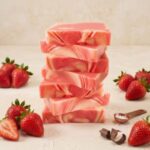 A 300x300 square professional food photograph of a tall, slightly leaning stack of seven rectangular fudge blocks. The fudge features a beautiful, artisanal swirl of vibrant strawberry pink and creamy off-white cheesecake colors. The stack is centered on a textured, light cream surface. Groups of fresh, ripe red strawberries with green leaves are arranged artfully on both the left and right sides of the base. In the lower-right foreground, a few dark chocolate curls and a small copper-toned measuring scoop filled with coarse sea salt crystals add to the gourmet presentation. The background is a soft, minimalist cream, creating a bright and cheerful aesthetic. High-key, natural lighting highlights the smooth, swirled texture of the fudge.
