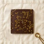A 300x300 square professional flat-lay photograph featuring a single, perfect square of Tennessee Whiskey milk chocolate. The chocolate has a rich, glossy brown finish and is perfectly centered on a light cream-colored linen napkin with visible hemstitch detailing. The aesthetic is clean and minimalist, with soft, even lighting that emphasizes the silky texture of the chocolate against the natural weave of the fabric. The composition is balanced and elegant, focusing entirely on the simple, indulgent quality of the chocolate.