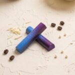 A 300x300 square professional food photograph featuring two cylindrical gourmet "The Moonshiner" truffle bars. The bars are arranged in an "X" shape, showcasing a stunning celestial aesthetic with a shimmering gradient of deep indigo blue and vibrant violet purple, speckled with fine silver dust to resemble a night sky. The text "THE MOONSHINER" is centered at the bottom in a bold, dark chocolate-toned serif font. The bars rest on a heavily textured, off-white cream surface. Decorative elements include scattered roasted coffee beans, delicate white chocolate shavings, and small white pearls. The lighting is soft and artistic, highlighting the iridescent "galaxy" finish of the truffles for a premium, magical look.