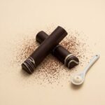 A high-resolution product photograph from an overhead angle, featuring two crossed cylindrical gourmet chocolate truffle bars on a cream-colored textured plaster surface. The bars are made of dark chocolate with internal white and green swirls, centrally placed on the plaster surface. Surrounding them are the same carefully curated aesthetic items from image_280.png: scattered dried jasmine flower buds on the left, a single roasted coffee bean and jasmine bud between the bars, and the two copper measuring spoons in the upper right. The gold serif text 'CHOCOLATE MINT' is retained below the bars. The scene is well-lit with soft, directional light, highlighting the textures of the bars, props, and plaster. The entire composition is balanced and has a curated, sophisticated feel.