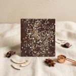 A 300x300 square professional food photograph of a square milk chocolate bar. The top of the bar is generously sprinkled with a mix of bright white shredded coconut and golden-brown toasted coconut flakes. The bar is positioned upright against a soft, draped cream-colored linen fabric. In the foreground, fresh coconut slices, dark chocolate curls, and star anise are artfully scattered. To the right, a small copper-toned measuring scoop holds toasted coconut bits. The background is a minimalist cream wall with subtle, curved textures, creating a warm, artisanal, and tropical aesthetic. Natural light emphasizes the contrasting textures of the smooth chocolate and the crunchy coconut.