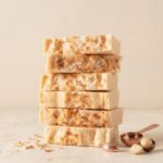 A close-up photograph of six squares of white coconut macadamia nut fudge stacked in a vertical column. Each piece has distinct wavy textures along the front and is topped with white and toasted coconut shavings. The stack is centered on a light cream-colored textured plaster surface against a seamless cream background. At the base of the stack are scattered toasted coconut shavings, two whole macadamia nuts (one shelled and broken), a copper-toned measuring scoop, and a small copper-colored cocoa bean charm. Soft, natural lighting.
