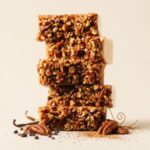 A tall, 300x300 square image featuring a stack of five thick white chocolate fudge slices. Each slice is densely topped with a thick layer of chopped, toasted pecans. The fudge stack sits on a light cream surface. At the base, whole pecans, vanilla beans, and cacao nibs are artfully arranged alongside a delicate gold wire accent and a dusting of cinnamon. The background is a soft, solid cream color, creating a warm and elegant professional food photograph.