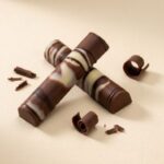 A 300x300 square professional food photograph featuring two cylindrical gourmet "Milk Chocolate Peanut Butter" truffle bars. The bars are made of rich milk chocolate and are decorated with an artistic dark chocolate drizzle pattern. They are arranged in an "X" shape on a soft, cream-colored woven linen surface. The scene is elegantly accessorized with small chocolate curls, whole roasted coffee beans, dried vanilla bean pods, jasmine flower buds, and a dust of edible gold. The text "MILK CHOCOLATE PEANUT BUTTER" is centered at the bottom in a bold, dark brown serif font. The lighting is soft and natural, emphasizing the textures for a high-end, artisanal aesthetic.