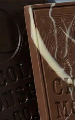 vertical, close-up shot showing the corner and side of two artisanal chocolate bars. The bar in the foreground is milk chocolate, featuring an elegant, embossed circular filigree design and the partially visible words "CHOCOLATE MOONSHINE." It is decorated with a diagonal, marbled swirl of white chocolate. Tucked behind it is a darker, plain chocolate bar with similar embossed lettering. The lighting is soft, highlighting the smooth texture and crisp edges of the chocolate.