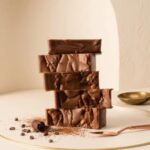 Five thick slices of artisanal chocolate stout fudge are stacked precisely on a white, gold-rimmed platform. The fudge has a rich, dark brown color with a naturally textured, rustic surface. Decorative elements surround the base, including a chocolate curl, a fine dusting of cocoa powder, and scattered cacao nibs. A rose-gold serving tool and a small brass bowl are positioned nearby on the cream surface. The background features a soft, arched cream wall, creating a minimalist and sophisticated product showcase.