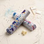 A 300x300 square professional food photograph featuring two cylindrical gourmet Birthday Cake truffle bars. The bars are arranged in an "X" shape and feature a white chocolate coating decorated with a vibrant splatter of blue, purple, pink, and yellow dots, mimicking birthday sprinkles. The text "BIRTHDAY CAKE" is centered at the bottom in a refined gold serif font. The bars rest on a textured, light cream-colored surface. Festive accents include small silver sugar pearls, a single white chocolate curl, light blue decorative twine, and two small white and gold flags on toothpicks in the background. The aesthetic is bright, celebratory, and artisanal.