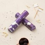 A high-resolution, overhead photograph showing two gourmet, galaxy-themed chocolate truffle bars crossed over each other. The bars are coated in dark purple chocolate and feature a striking "milky way" splatter pattern of white and lighter purple speckles. Large, irregular white chocolate patches decorate the bars. The bars rest on a textured, off-white (cream-colored) plaster or stone surface. The surrounding area is decorated with small, silver edible sugar pearls, white chocolate curls, a copper-toned measuring cup filled with fresh blackberries, and a pair of small, gold and white decorative flags on toothpicks. The lighting is soft and even, highlighting the unique textures.