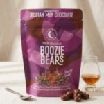 boozie-bears A 300x300 square professional flat-lay photograph featuring a single, perfect square of Tennessee Whiskey milk chocolate. The chocolate has a rich, glossy brown finish and is perfectly centered on a light cream-colored linen napkin with visible hemstitch detailing. The aesthetic is clean and minimalist, with soft, even lighting that emphasizes the silky texture of the chocolate against the natural weave of the fabric. The composition is balanced and elegant, focusing entirely on the simple, indulgent quality of the chocolate.