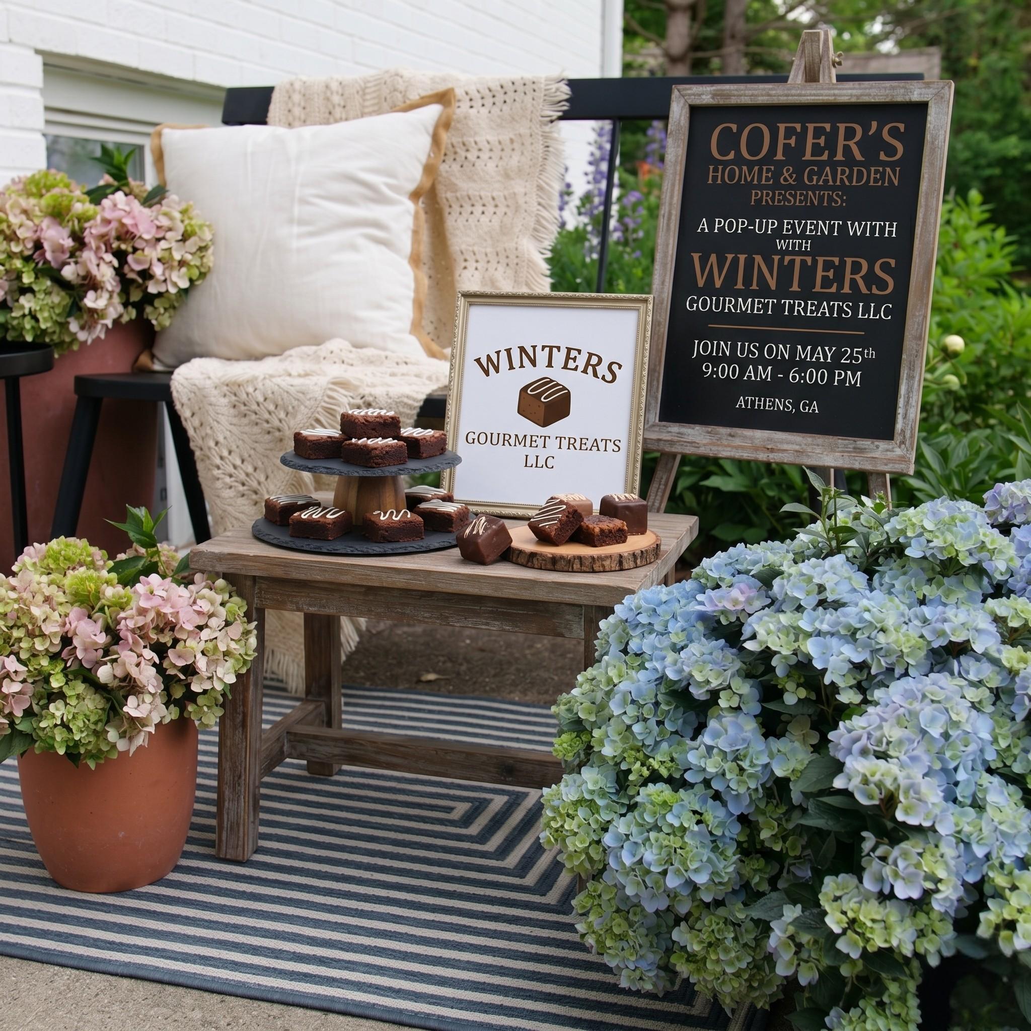 An inviting outdoor display setup on a striped rug featuring gourmet brownies on a wooden table. The arrangement is surrounded by large blue and pink hydrangea flowers. Two signs present event details: one framed sign shows the 'Winters Gourmet Treats LLC' logo, and a large easel chalkboard reads 'Cofer's Home & Garden Presents: A Pop-Up Event with Winters Gourmet Treats LLC, Join Us On May 25th 9:00 AM - 6:00 PM, Athens, GA.' A rustic bench with a pillow and blanket sits in the background.