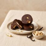 dark-chocolate-cashew-cups A 300x300 square professional flat-lay photograph featuring a square Belgian milk chocolate bar. The surface of the chocolate is studded with whole and crushed vibrant green roasted pistachios. The bar is centered on a light, textured cream-colored surface. To the left, small clusters of whole pistachios, dark chocolate curls, and star anise are artfully arranged. To the right, two elegant gold-toned measuring scoops rest on the surface, one containing coarse sea salt crystals. The lighting is soft and natural, emphasizing the rich, smooth texture of the chocolate and the crunchy, colorful pistachio topping for a sophisticated, artisanal look.