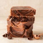 A central, eye-level photograph of five thick, square blocks of rich, dark chocolate coffee fudge stacked vertically. The fudge has a beautiful, smooth marbled pattern of deep cocoa brown and warm caramel tones throughout. The stack is centered on a light beige textured plaster-like surface. To the lower left, a single whole roasted coffee bean, a small dark chocolate curl, and a scattered smaller piece of chocolate rest. To the right of the stack, a polished, copper-toned measuring scoop holds a small amount of coffee. Two more small dark chocolate curls are near the base of the stack to the right. The background is a soft, seamless light beige textured surface. The lighting is soft and directional, highlighting the rich textures and colors.