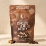 espresso-bean-medley A 300x300 square professional flat-lay photograph featuring a square Belgian milk chocolate bar. The surface of the chocolate is studded with whole and crushed vibrant green roasted pistachios. The bar is centered on a light, textured cream-colored surface. To the left, small clusters of whole pistachios, dark chocolate curls, and star anise are artfully arranged. To the right, two elegant gold-toned measuring scoops rest on the surface, one containing coarse sea salt crystals. The lighting is soft and natural, emphasizing the rich, smooth texture of the chocolate and the crunchy, colorful pistachio topping for a sophisticated, artisanal look.