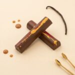 A professional food photograph featuring two crossed cylindrical gourmet truffle bars. The bars have a deep burgundy finish with subtle green undertones, presented on a cream-colored textured plaster surface. The scene is elegantly curated with scattered roasted coffee beans, small dried natural flower petals, and two tiny polished copper measuring spoons in the corners. Centered below the chocolate bars, the text "HOT MESS" is written in a sophisticated brown serif font.