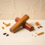 A 300x300 square professional food photograph featuring two cylindrical gourmet Mimosa truffle bars. The bars are arranged in an "X" shape and feature a striking ombre finish transitioning from a reddish-orange to a bright yellow. The text "MIMOSA" is centered at the bottom in a bold, copper-toned serif font. The bars rest on a textured, light cream-colored surface. Scattered around the base are pieces of dried orange peel, small dried fruit pieces, whole roasted cacao nibs, and delicate white decorative pearls. The lighting is soft and elegant, creating a high-end, artisanal aesthetic with a tropical, summery feel.