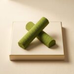 A professional food photograph featuring two crossed cylindrical gourmet truffle bars. The bars have a deep burgundy finish with subtle green undertones, presented on a cream-colored textured plaster surface. The scene is elegantly curated with scattered roasted coffee beans, small dried natural flower petals, and two tiny polished copper measuring spoons in the corners. Centered below the chocolate bars, the text "HOT MESS" is written in a sophisticated brown serif font.