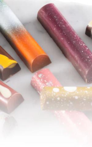 collection of slender, rounded artisanal chocolate bars displayed on a light marble background. Each bar features a unique, vibrant design: one has a gradient of orange and silver, another is deep plum with white speckles, and others show splashes of yellow and pink. The chocolates have a smooth, glossy finish, suggesting a high-end, handcrafted quality. The bottom of the image fades into a soft white mist