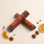 A 300x300 square professional food photograph featuring two cylindrical gourmet Orange Bourbon truffle bars. The bars are arranged in an "X" shape and feature a warm, copper-orange metallic finish decorated with horizontal dark chocolate stripes. The bars rest on a textured, light cream-colored surface. The composition is accented with two dried orange slices, several whole star anise pods, and scattered roasted cocoa nibs. The lighting is soft and warm, creating a premium, artisanal, and autumnal aesthetic.