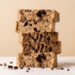 A vertical stack of five thick, rectangular slices of peanut butter fudge mixed with crushed chocolate sandwich cookies. The fudge is a light tan color, densely packed with dark cookie chunks and fine crumbs throughout. The stack sits on a textured white surface, with chocolate cookie pieces and cacao nibs artistically scattered around the base. The background is a solid, warm cream color, providing a clean and appetizing presentation.