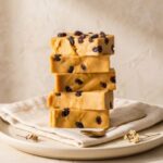 A 300x300 square professional food photograph of a tall stack of five rectangular Penuche fudge blocks. The fudge is a uniform, warm brown sugar and caramel color with a matte, creamy finish and subtle artisanal ripples on the sides. The stack is centered on a light, textured cream-colored surface. In the lower-left, several dark vanilla bean pods and a few small chocolate shavings are artfully arranged. To the right, additional dark chocolate curls rest near the base. The background is a minimalist, textured cream color, creating a warm, sophisticated aesthetic. Soft lighting highlights the dense, smooth texture of the brown sugar fudge.