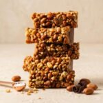 A 300x300 square professional food photograph featuring a tall stack of five rectangular fudge blocks inspired by Snickers. The front of each slice is densely packed with a thick layer of roasted whole and chopped peanuts. The stack is centered on a light, textured cream-colored surface. In the lower-left, a small copper measuring scoop holds coarse sea salt next to a scattered whole peanut. To the right, dark chocolate curls and additional roasted peanuts are artfully arranged. The background is a soft, minimalist cream, creating a warm and sophisticated aesthetic. Bright, natural lighting emphasizes the crunchy, salty-sweet texture of the peanut topping.