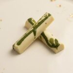 A professional food photograph featuring two crossed cylindrical gourmet truffle bars. The bars have a deep burgundy finish with subtle green undertones, presented on a cream-colored textured plaster surface. The scene is elegantly curated with scattered roasted coffee beans, small dried natural flower petals, and two tiny polished copper measuring spoons in the corners. Centered below the chocolate bars, the text "HOT MESS" is written in a sophisticated brown serif font.